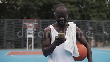 年轻肌肉发达，微笑着的美国黑人，一只手拿着球，一只手拿着智能手机